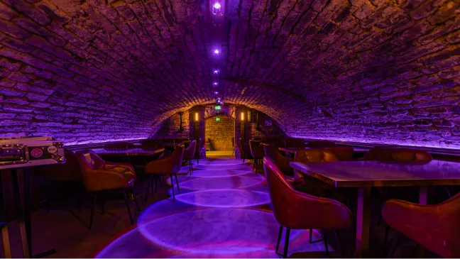 Das Kellergewölbe in violettem Licht mit Tischen und Stühlen an der Steinwand.